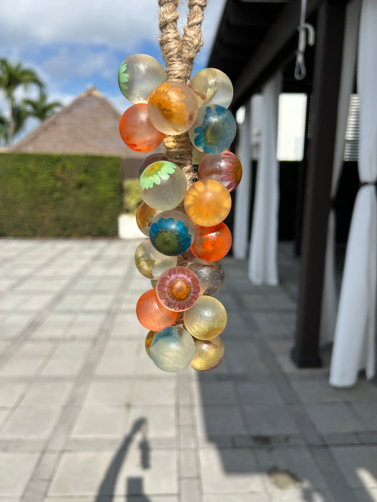 A grape cluster soap featuring translucent spheres with embedded colorful flowers in shades of orange, blue, yellow, and green hanging outdoors by a rope loop, with a blurred patio in the background