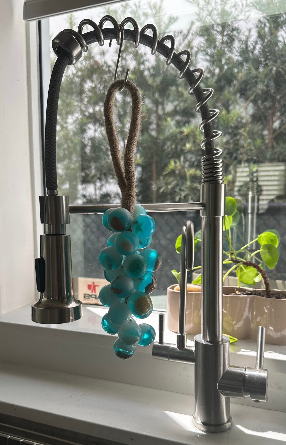 A grape cluster style soap made of turquoise blue spheres hanging by a brown fiber loop from a modern stainless steel kitchen faucet. The scene is illuminated by natural light coming through a window, with a green potted plant visible on the sill behind the sink.