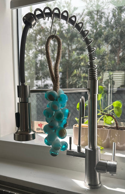 A grape cluster style soap made of turquoise blue spheres hanging by a brown fiber loop from a modern stainless steel kitchen faucet. The scene is illuminated by natural light coming through a window, with a green potted plant visible on the sill behind the sink.