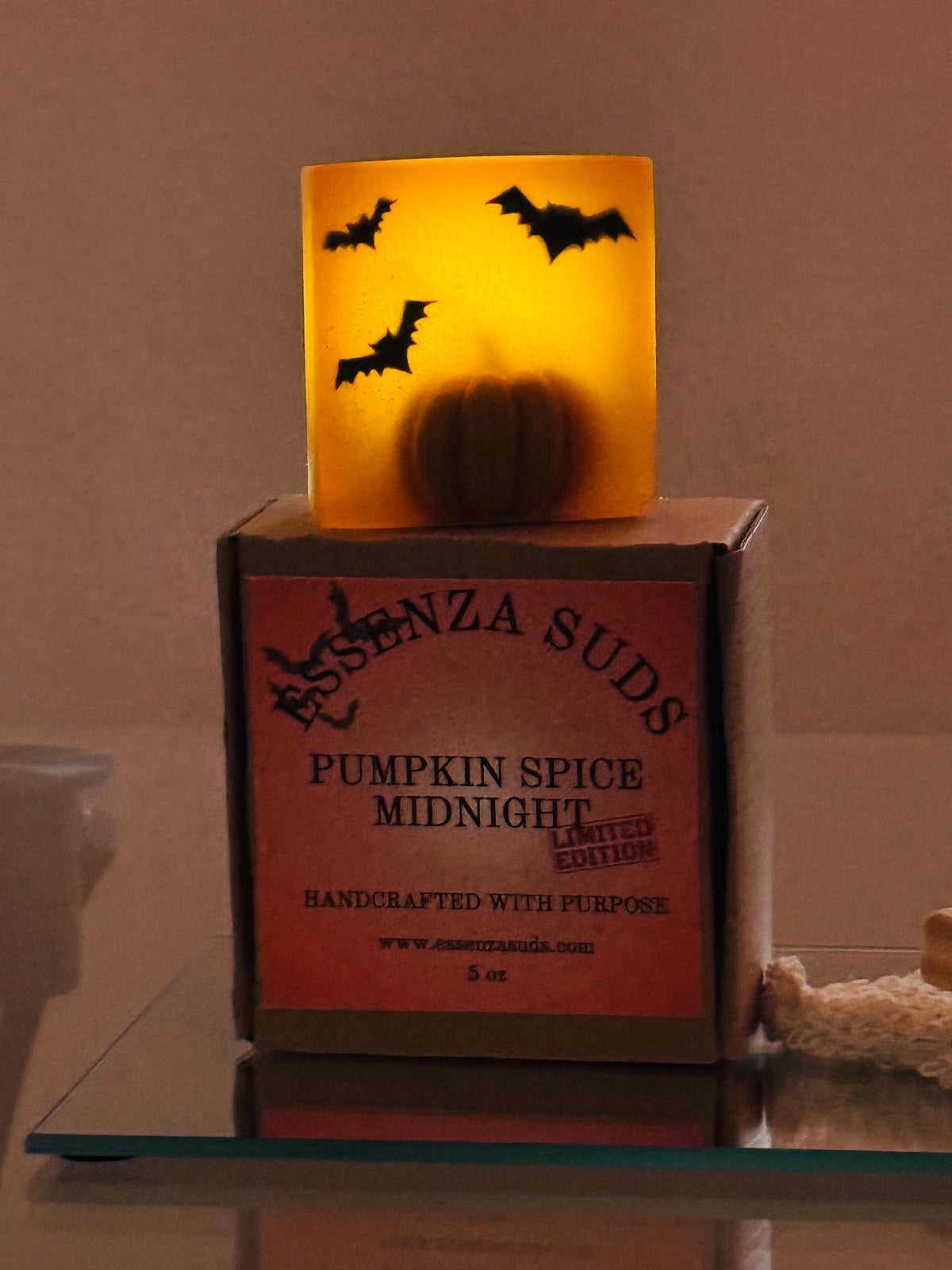 A square bar of ESSENZA SUDS Pumpkin Spice Midnight soap with a yellow and orange gradient, embedded pumpkin, and bat silhouettes, sitting on its Kraft paper packaging box.