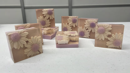Several rectangular decorative soap bars with large daisy flower designs are arranged on a light speckled surface. The soaps have a warm beige base with cream flowers and soft purple centers, and some are standing upright while others are propped open to show the layered interior.