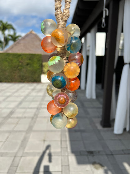 A grape cluster soap featuring translucent spheres with embedded colorful flowers in shades of orange, blue, yellow, and green hanging outdoors by a rope loop, with a blurred patio in the background