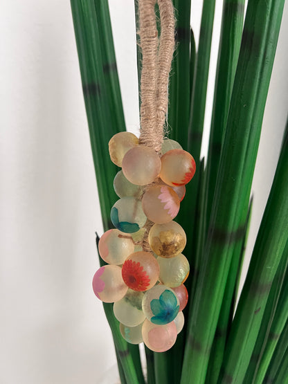 A close-up of a decorative floral arrangement featuring a grape cluster soap of translucent , round grapes soap in pastel pink, peach, cream, and teal, attached to a beige twine wrap and set against green faux pine needles on a white background.