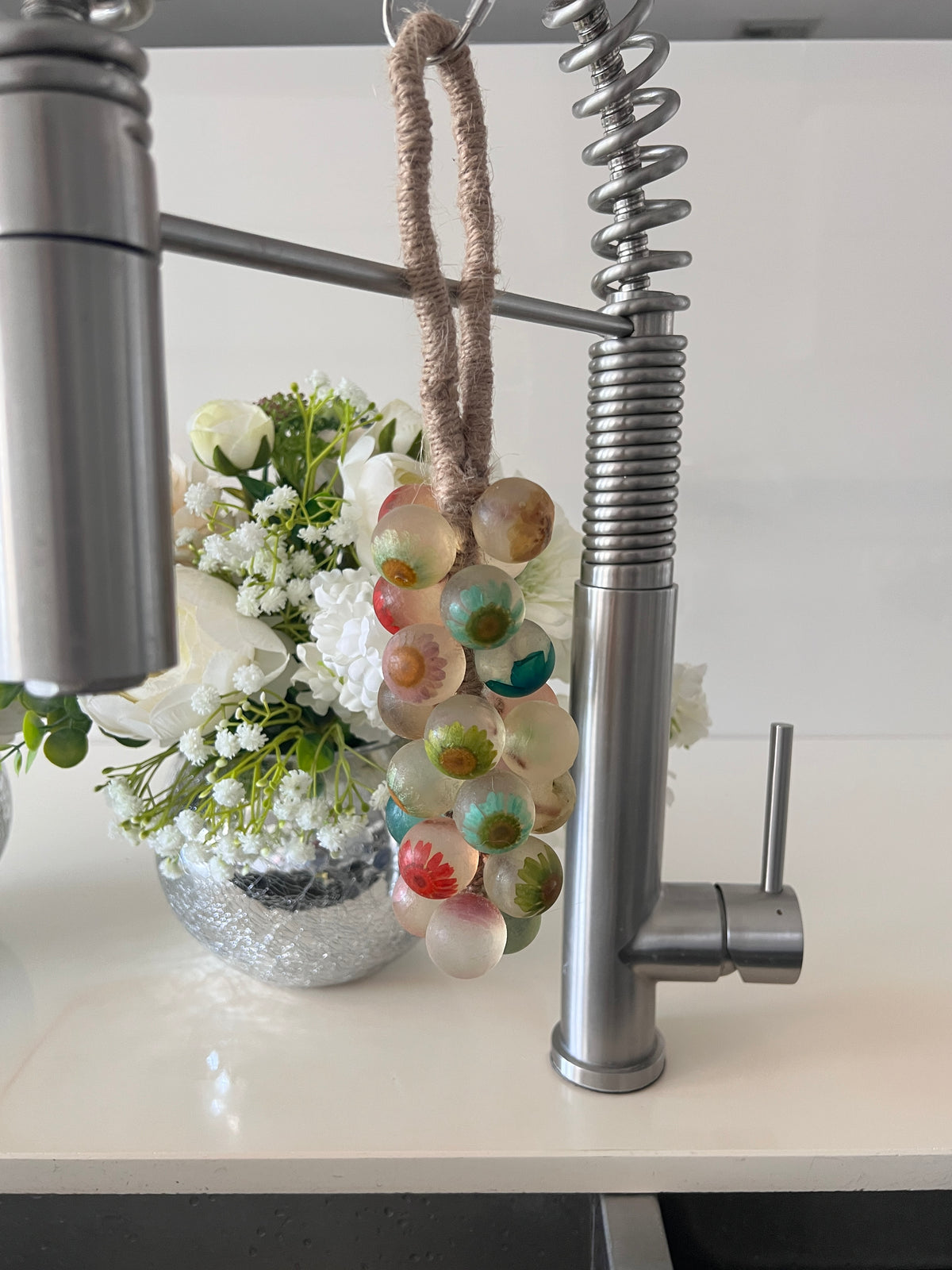 A grape cluster style soap hanging from a kitchen faucet by a brown rope loop. The soap is made of clear spheres, each embedding a colorful flower in shades of yellow, pink, turquoise, green, red, and blue