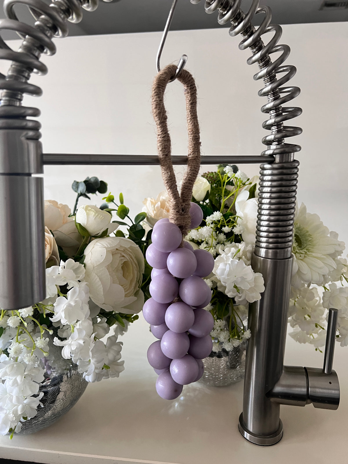 A grape cluster style soap hanging from a kitchen faucet by a brown rope loop. The soap is made of lilac spheres.