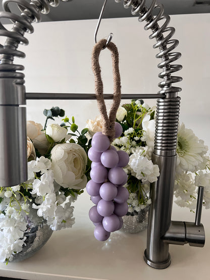 A grape cluster style soap hanging from a kitchen faucet by a brown rope loop. The soap is made of lilac spheres.