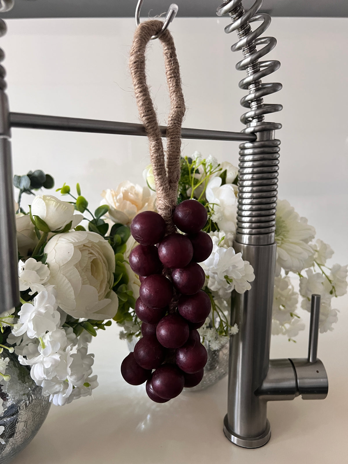 A grape cluster style soap hanging from a kitchen faucet by a brown rope loop. The soap is made of wine red spheres.