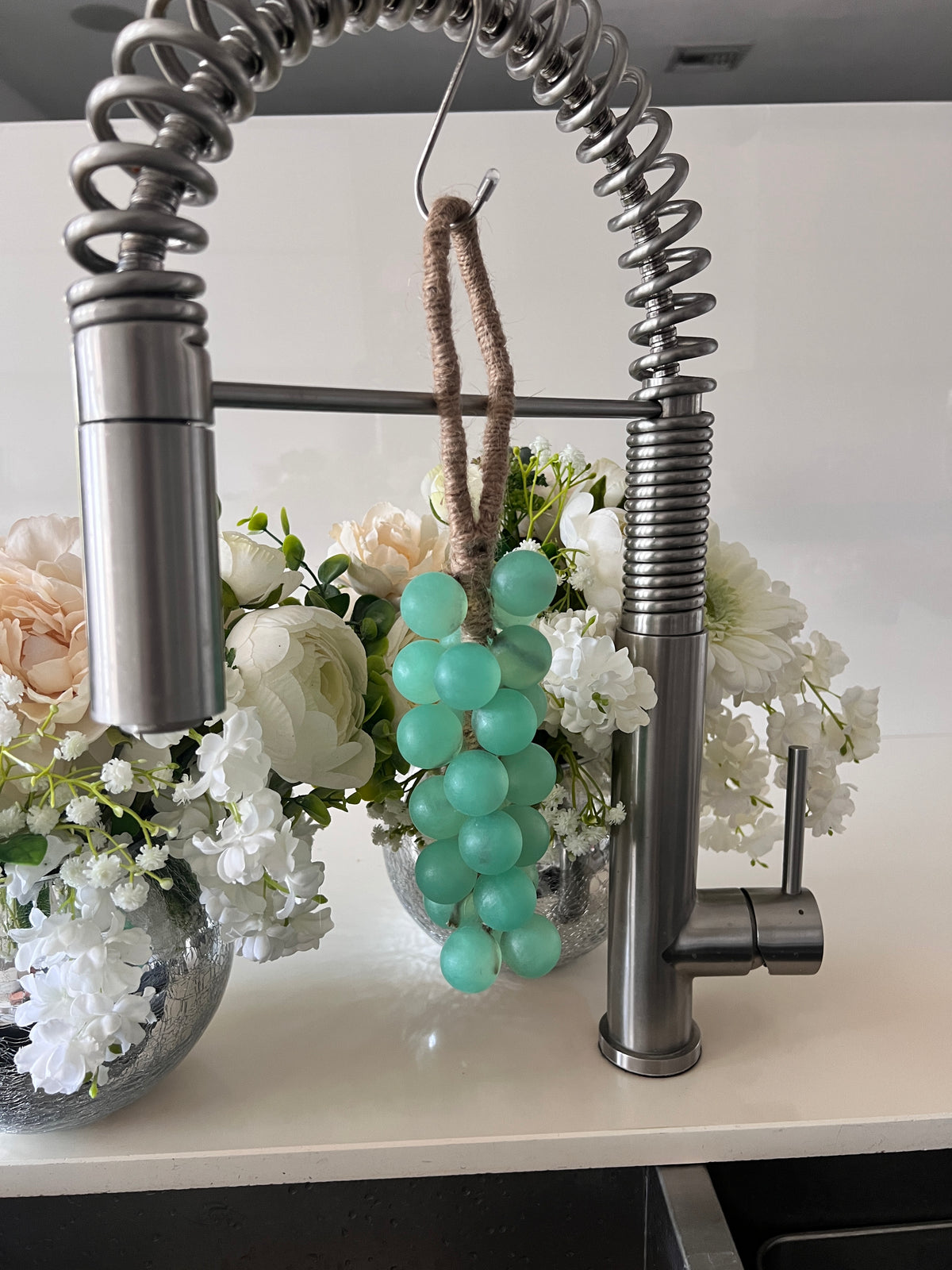 A grape cluster style soap hanging from a kitchen faucet by a brown rope loop. The soap is made of seafoam green spheres.