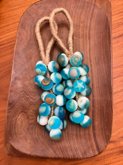 A grape cluster soap of round turquoise and white seashell beads on a tan cord, displayed on a dark wood serving board over a wooden table.