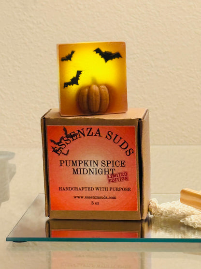 A boxed  labeled "Essenza Suds Pumpkin Spice Midnight" sits on a glass tray with a bar glowing pumpkin shaped soap displayed above the box. The soap bar packaging is orange and brown, and the lit candle shows a warm yellow-orange glow with black bat silhouettes.