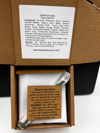 This image shows the thoughtful, cardboard packaging for our Palo Santo handmade soap by Essenza Suds., you'll see your soap snugly wrapped in protective white tissue paper. Also included and wrapped is a genuine Palo Santo smudging stick, accompanied by an informative card labeled "Enhance Your Ritual." This card explains how to burn the wood to cleanse your space's energy, transforming your bath time into a mindful, renewing experience.